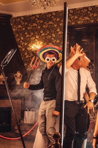 Dwie osoby pozują do zdjęcia w fotobudce: jedna w sombrero i okularach w kształcie kwiatów trzymająca plastikowe widły, druga w masce konia z krawatem, na tle dekoracyjnej ściany i ozdobnego...