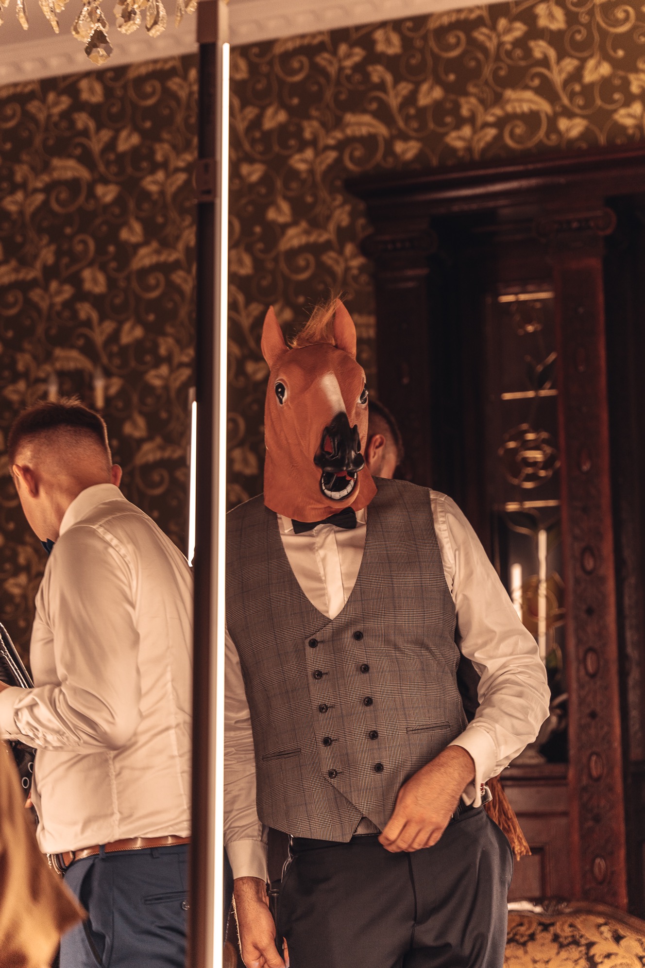 Osoba w masce konia i eleganckiej kamizelce pozuje w pomieszczeniu z dekoracyjną tapetą i oświetleniem LED.