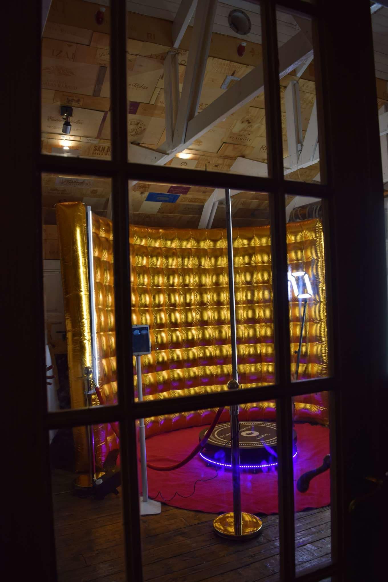 Widok przez okno na złotą dmuchaną fotobudkę z podświetlanym obrotowym podestem na czerwonym dywanie.