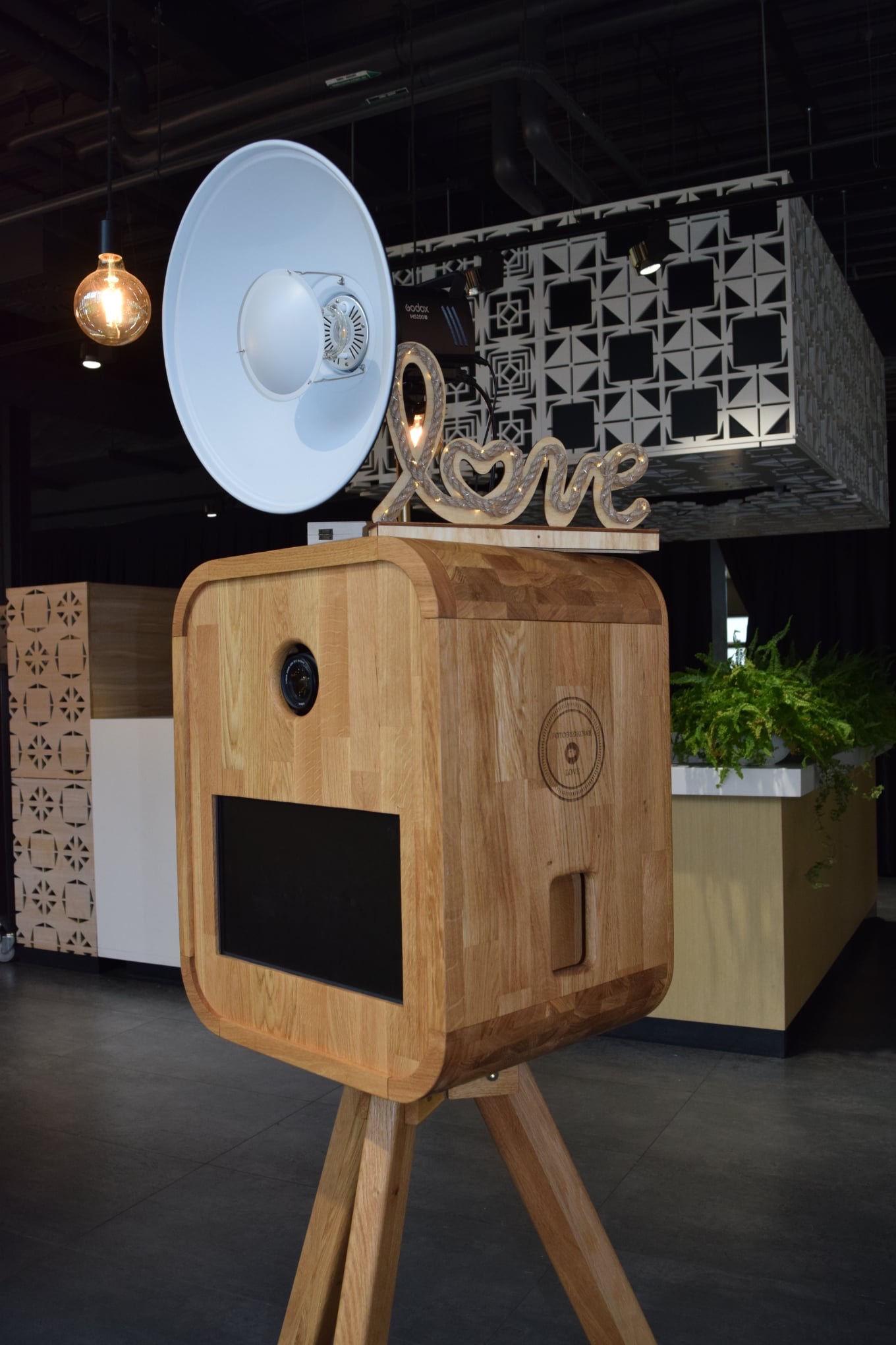 Stylowa fotobudka z drewna na trzech nogach, z napisem 'Love' i lampą błyskową w kształcie tuby. W tle dekoracje i oświetlenie eventowe.