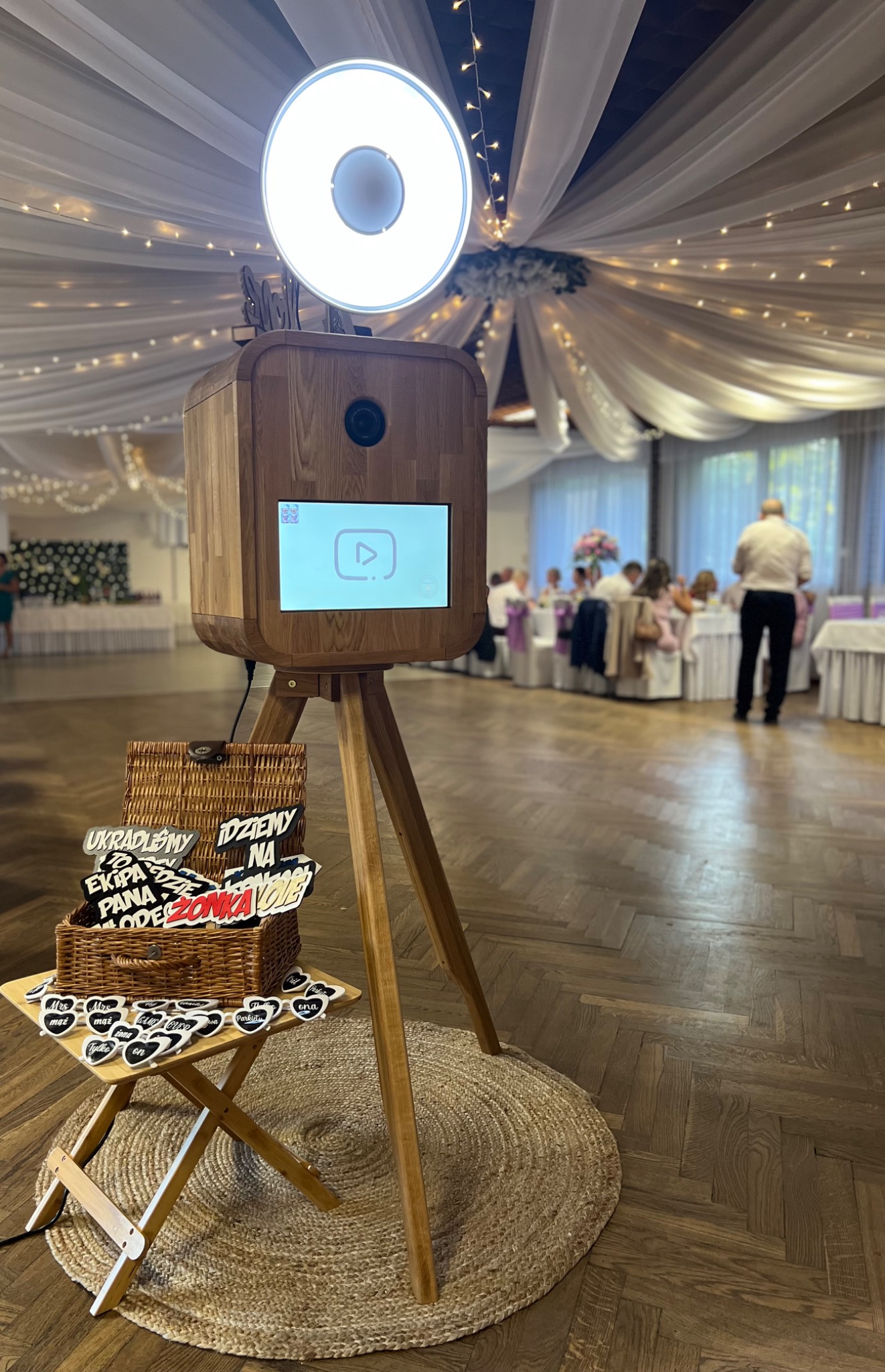Drewniana fotobudka na trójnogu z rekwizytami w koszyku, ustawiona na jutowym dywaniku podczas przyjęcia z gośćmi w tle.