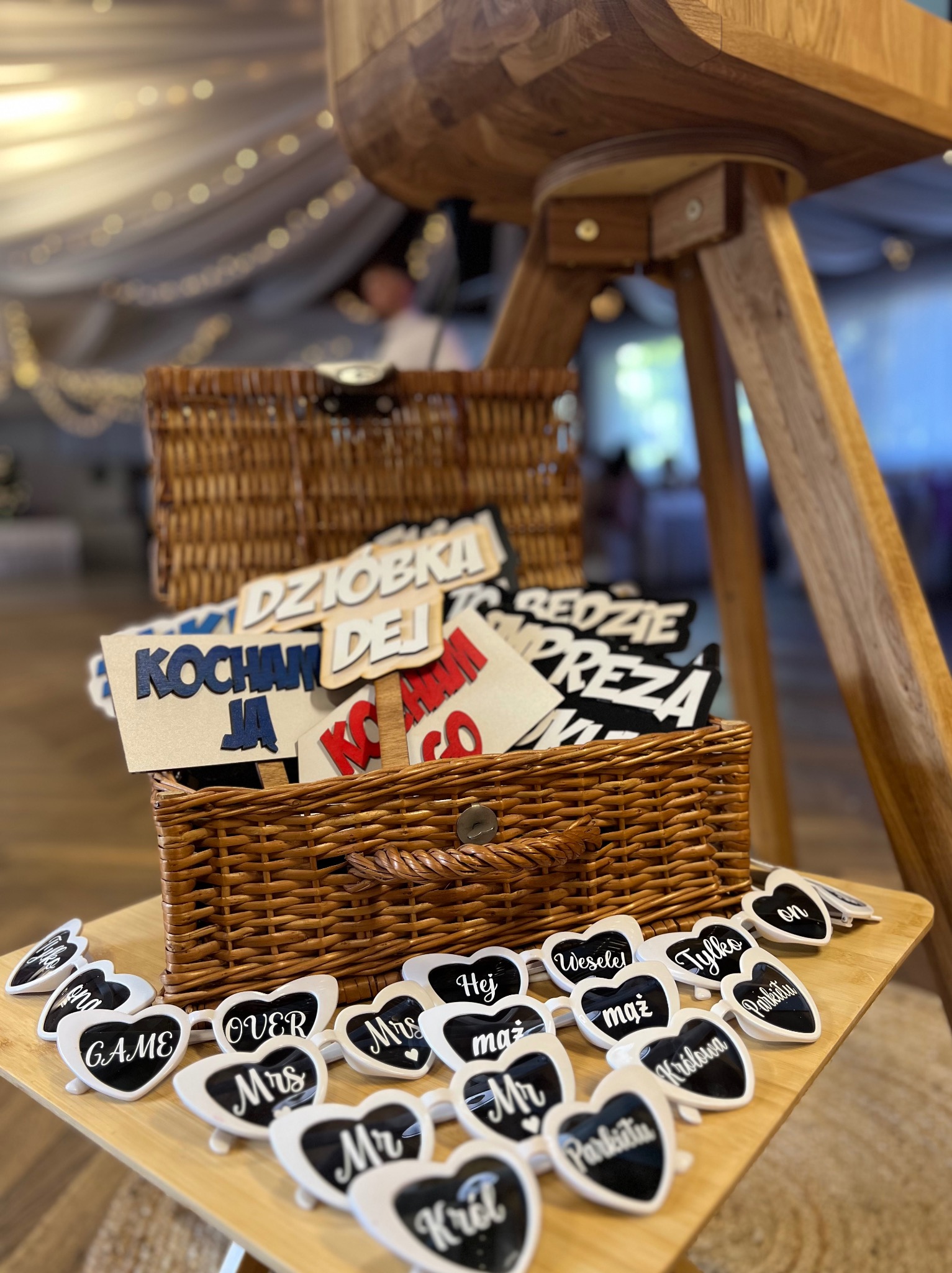 Rekwizyty do fotobudki: kosze z napisami i okulary na drewnianym stoliku, w tle sala bankietowa z dekoracjami świetlnymi.