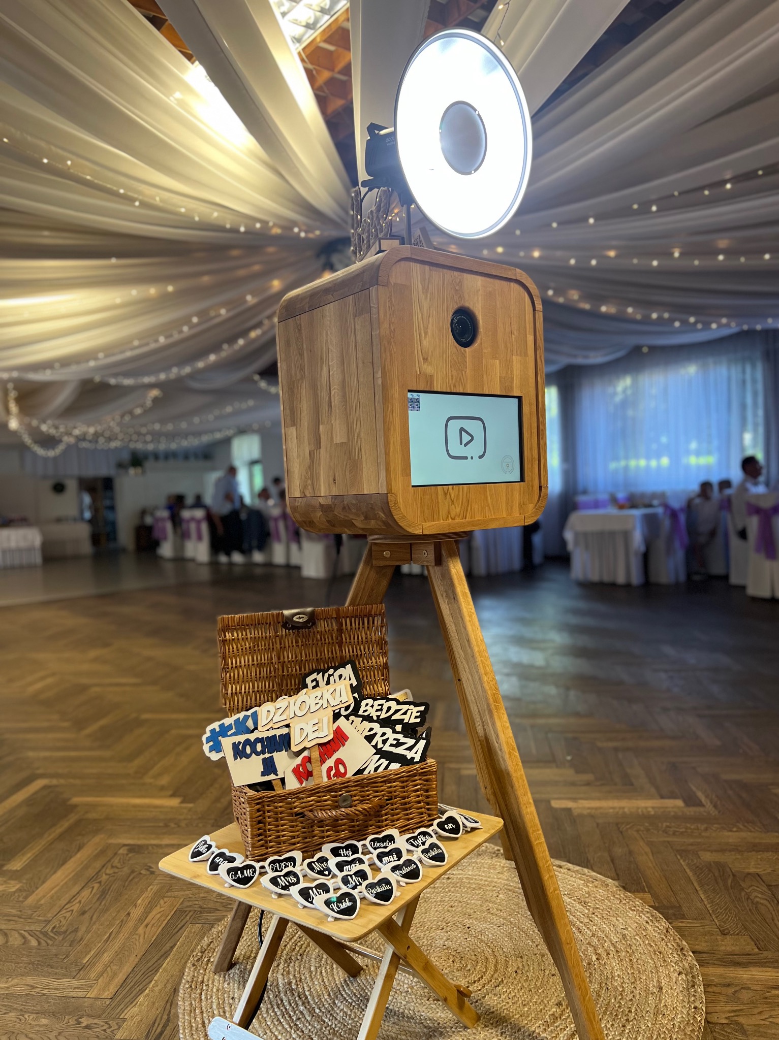 Drewniana fotobudka na weselu z rekwizytami: koszyk z napisami i okulary na stoliku. W tle sala z dekoracjami.