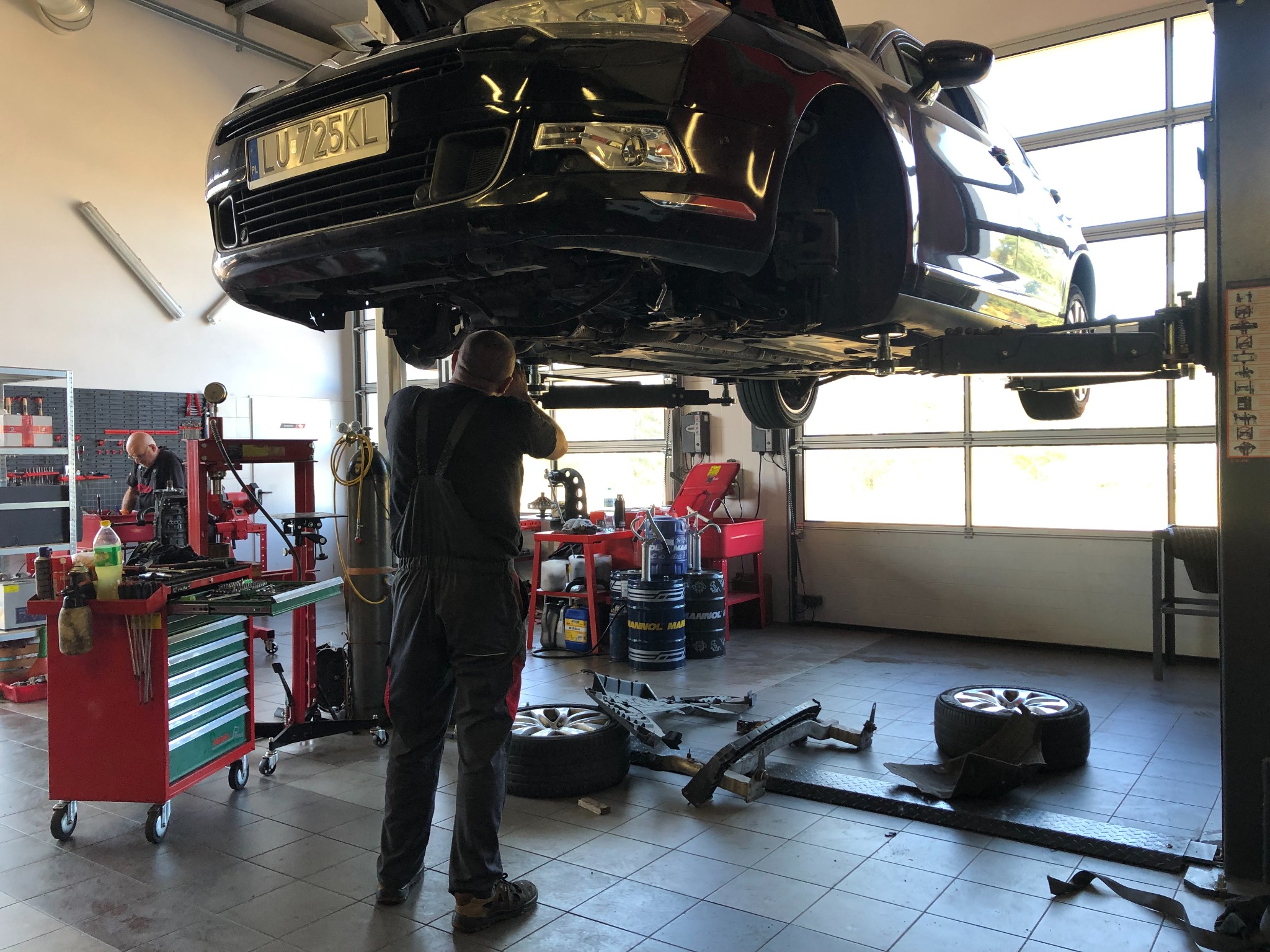 Czarny samochód na podnośniku w warsztacie samochodowym, mechanik w kombinezonie reguluje jego pozycję, w tle narzędzia i wyposażenie warsztatu.