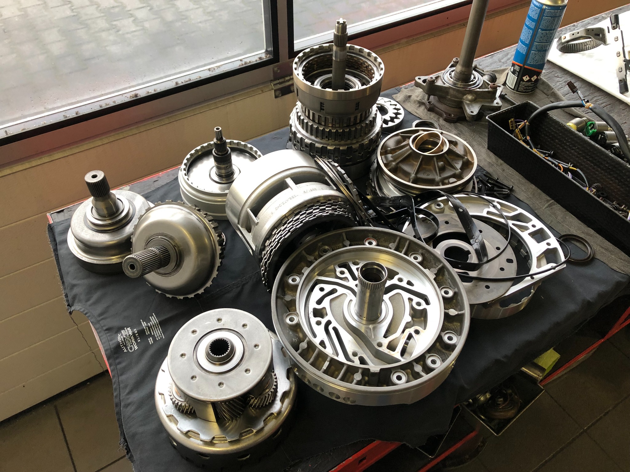 Rozłożona automatyczna skrzynia biegów na stole warsztatowym, widoczne różne elementy składowe, w tym przekładnie, tarcze cierne i wałki. Na stole widoczny również smar w sprayu i pojemnik...