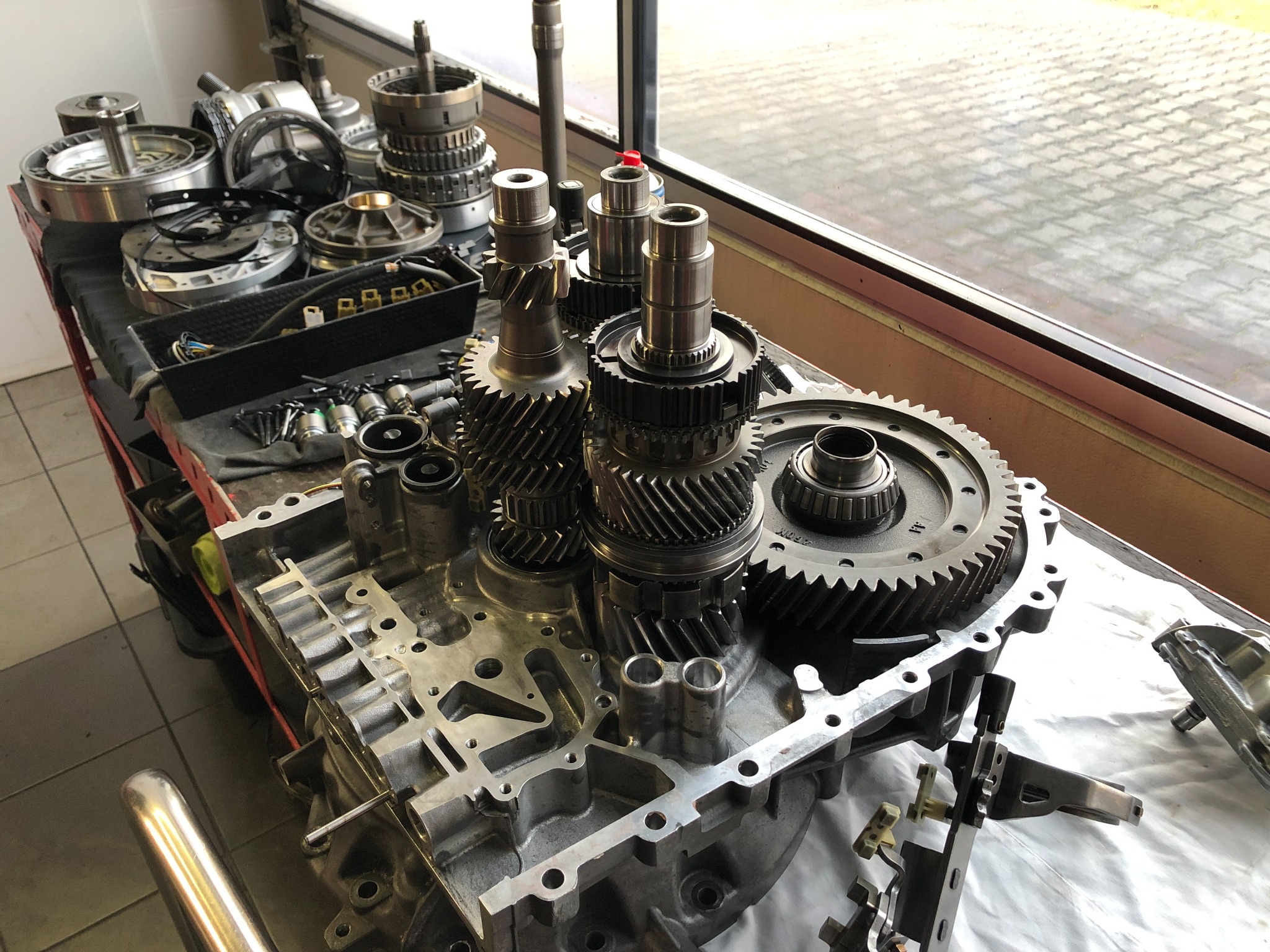 Rozłożona skrzynia biegów z widocznymi przekładniami, wałkami i obudową na warsztatowym stole w pobliżu okna.