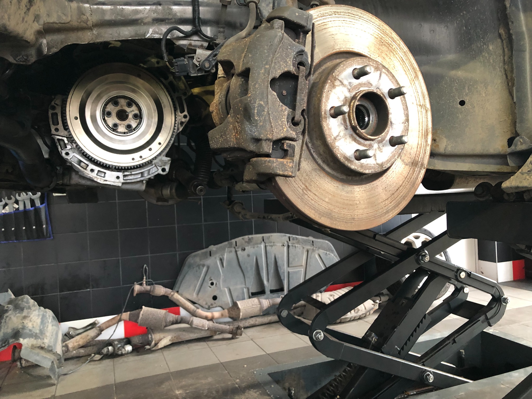 Widok podwozia samochodu na podnośniku, ukazujący tarczę hamulcową, zacisk, koło zamachowe oraz elementy układu wydechowego leżące na podłodze warsztatu.