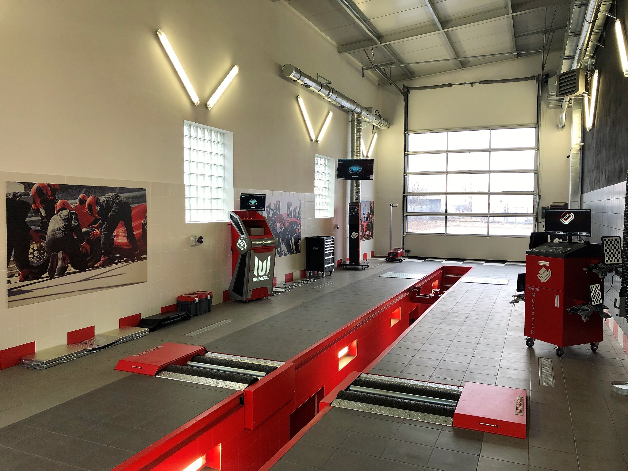 Wnętrze warsztatu samochodowego z kanałem diagnostycznym, urządzeniami do geometrii kół i plakatem przedstawiającym pracę mechaników przy bolidzie Formuły 1