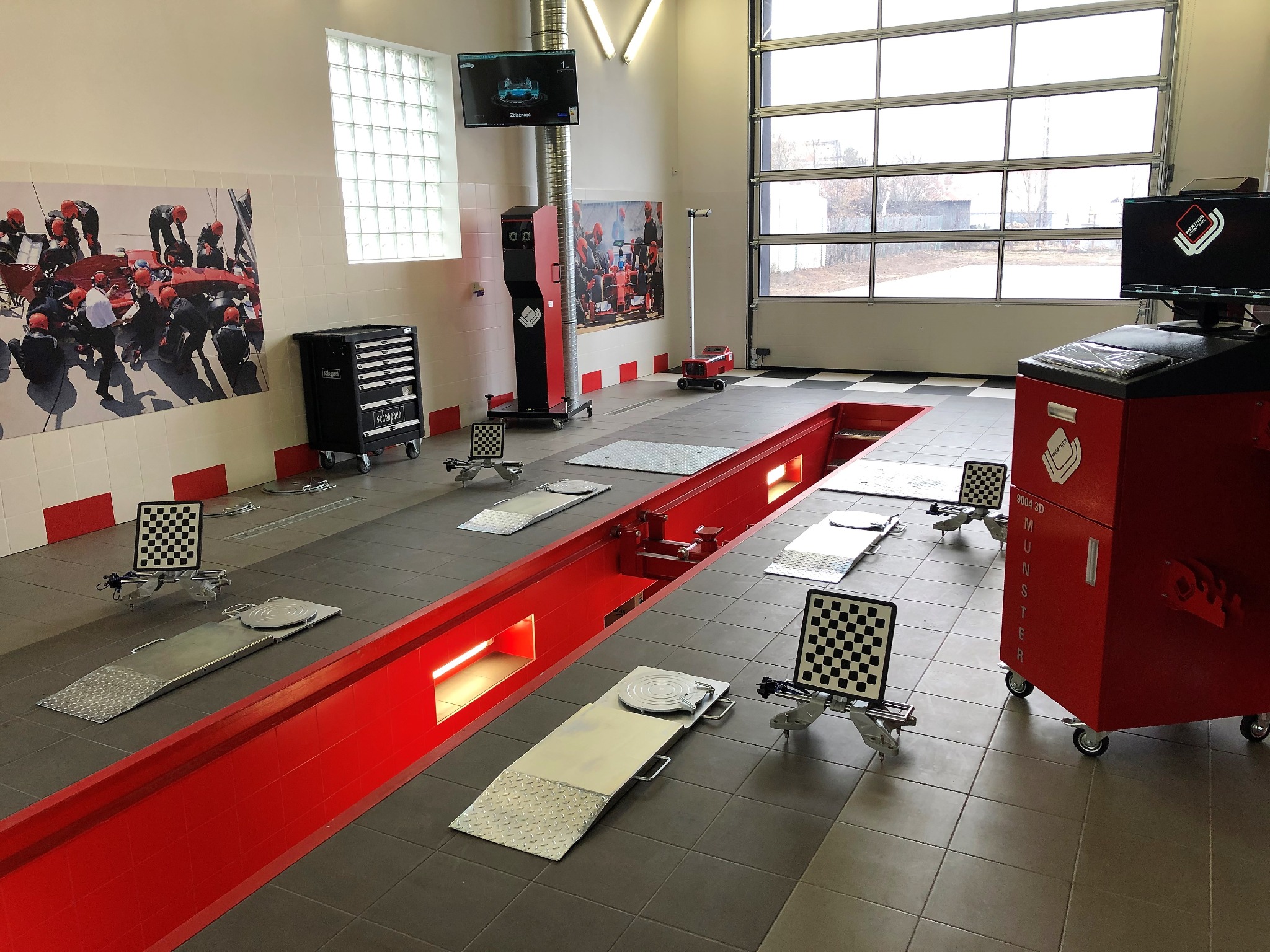 Wnętrze warsztatu samochodowego z kanałem diagnostycznym, stanowiskiem do geometrii kół i ścianą ozdobioną grafiką związaną z Formułą 1.