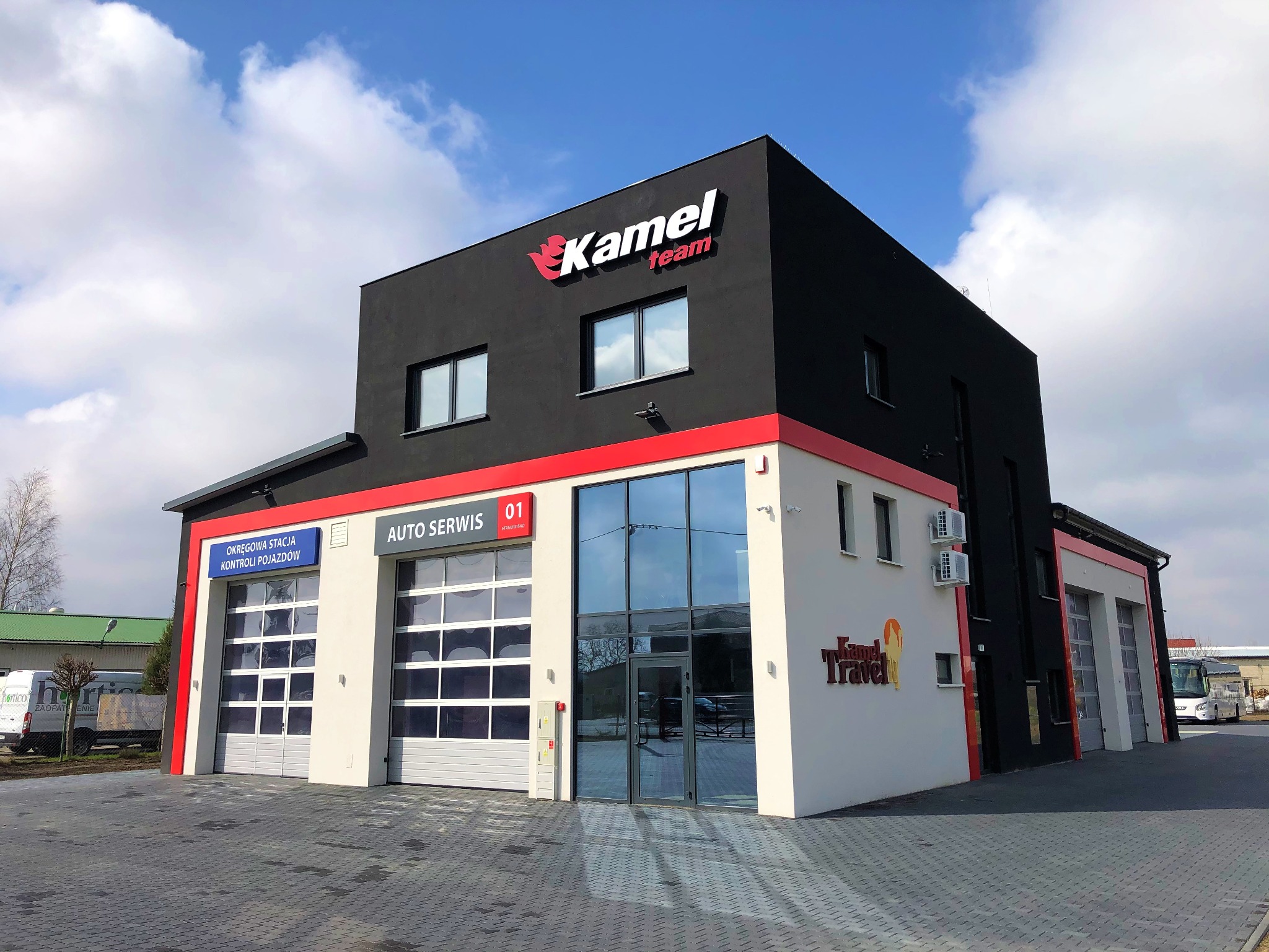 Dwukondygnacyjny budynek auto serwisu z bramami garażowymi, logo firmy 'Kamel Team' na czarnej elewacji i napisem 'Auto Serwis' nad wejściem, widok z zewnątrz w słoneczny dzień.