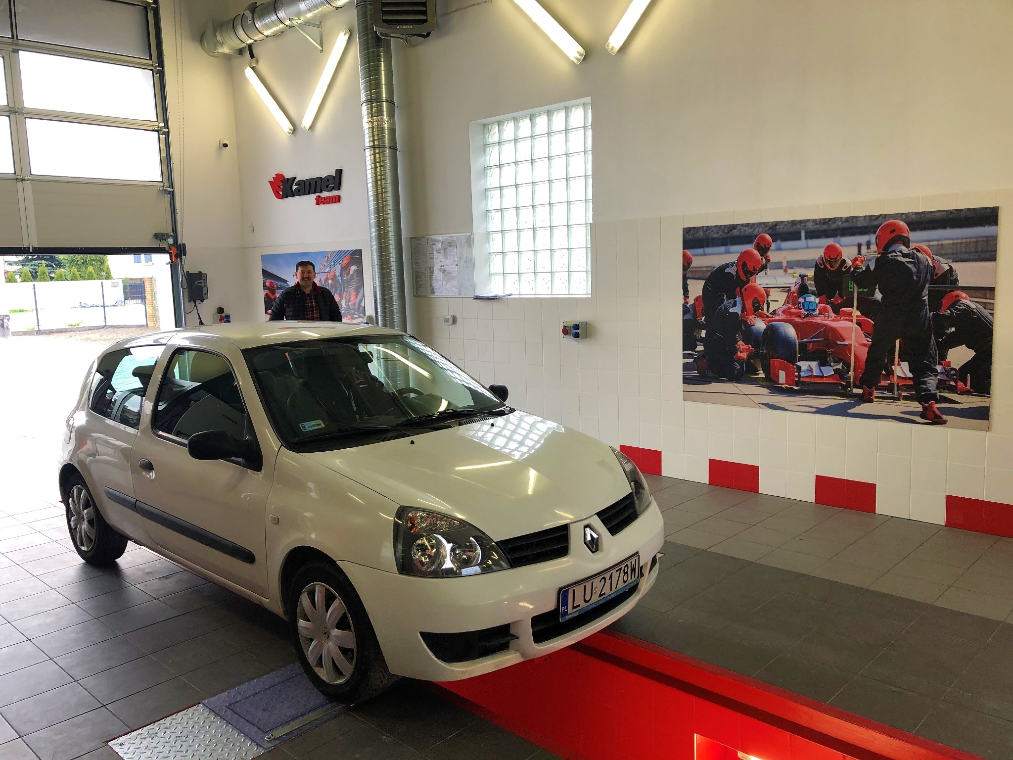 Białe Renault Clio na stanowisku diagnostycznym w jasnym warsztacie; w tle widoczny mechanik i plakat z zespołem Formuły 1.