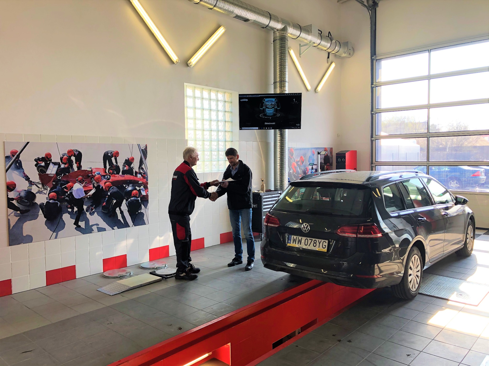 Wnętrze warsztatu samochodowego: mężczyźni w trakcie wymiany dokumentów obok podnośnika z czarnym kombi Volkswagen, na ścianie plakat z pit stopem Formuły 1 i monitor z diagnostyką pojazdu.