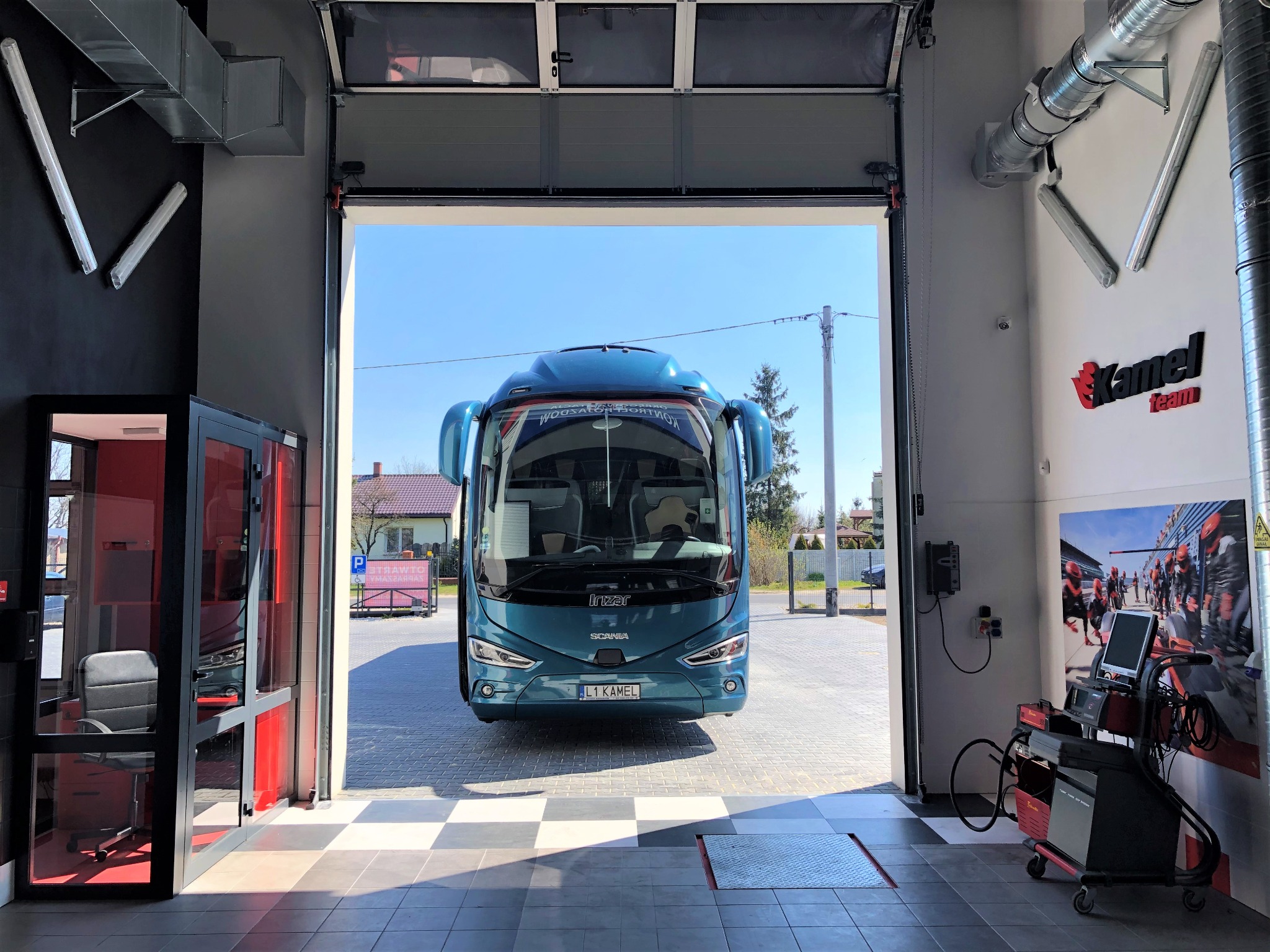Turkusowy autobus Scania wjeżdża do warsztatu przez otwartą bramę garażową, widoczny sprzęt diagnostyczny i logo firmy na ścianie.