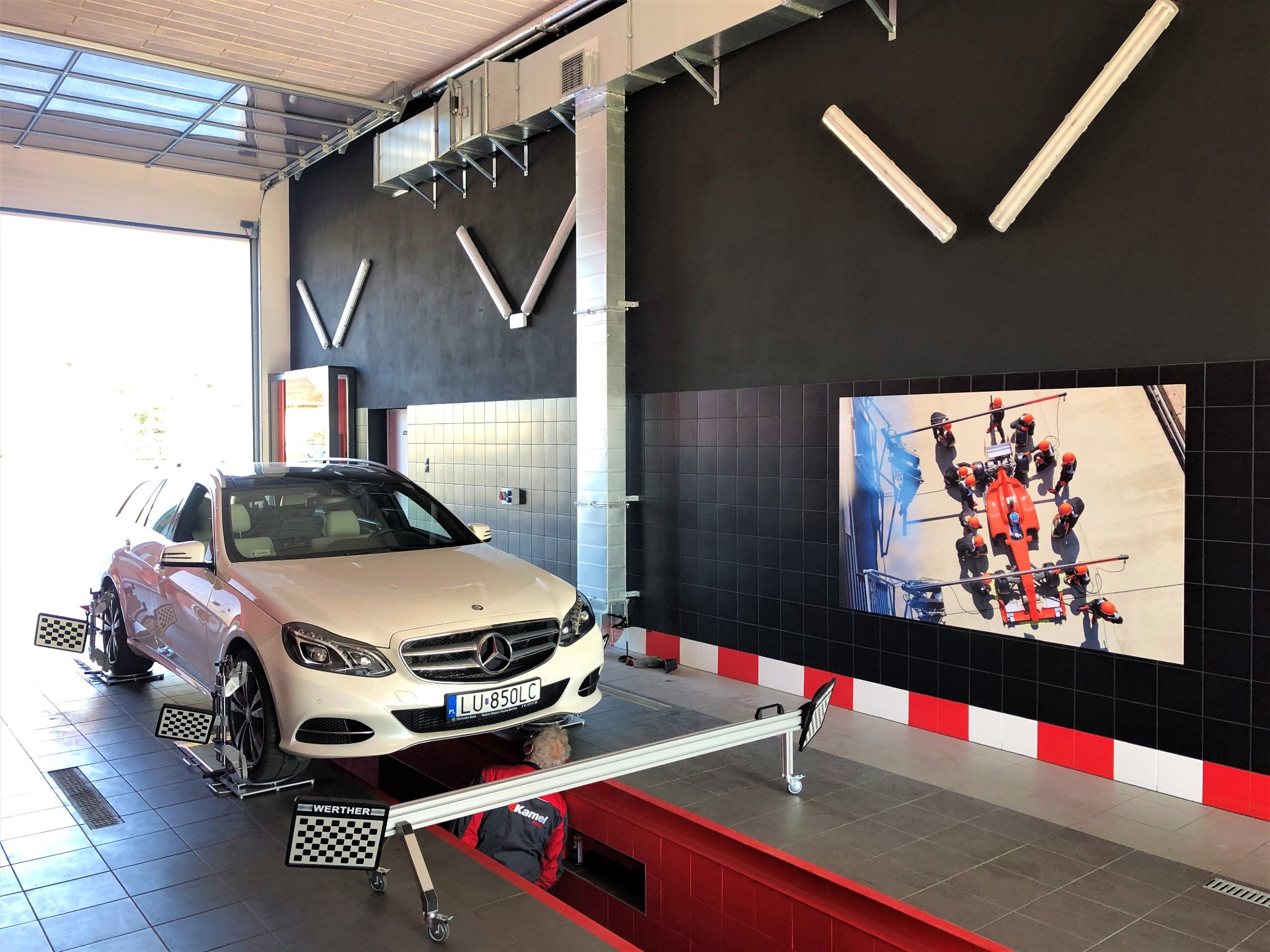 Biały Mercedes na stanowisku do geometrii kół w warsztacie, widoczny pracownik w kanale diagnostycznym, na ścianie zdjęcie bolidu Formuły 1.