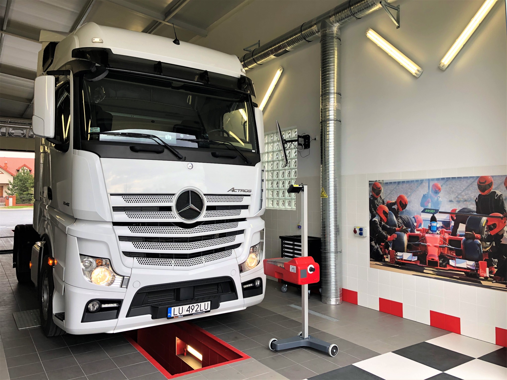 Biała ciężarówka marki Mercedes-Benz Actros na stanowisku diagnostycznym w warsztacie, z widocznym kanałem rewizyjnym i urządzeniem do ustawiania świateł, na ścianie plakat z bolidem Formuły 1.