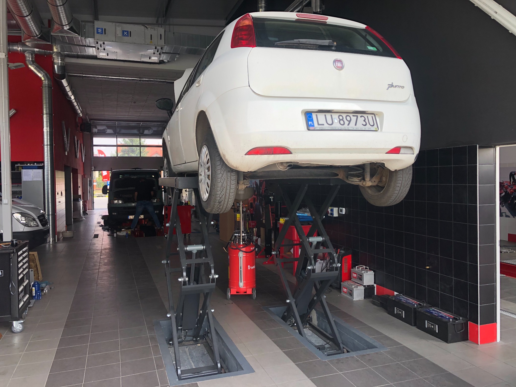 Biały samochód Fiat Punto podniesiony na podnośniku dwukolumnowym w warsztacie samochodowym, widoczna tablica rejestracyjna LU 8973U, w tle inne pojazdy i narzędzia.