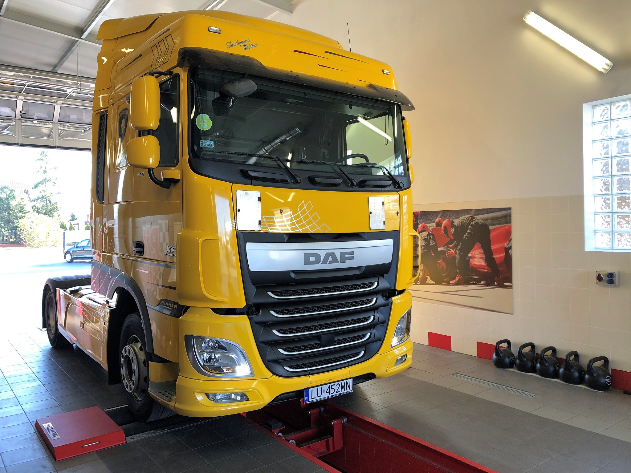 Żółty ciągnik siodłowy DAF na stanowisku diagnostycznym w warsztacie, widoczny kanał rewizyjny i podnośnik, w tle plakat z wyścigami samochodowymi i rząd odważników kettlebell.