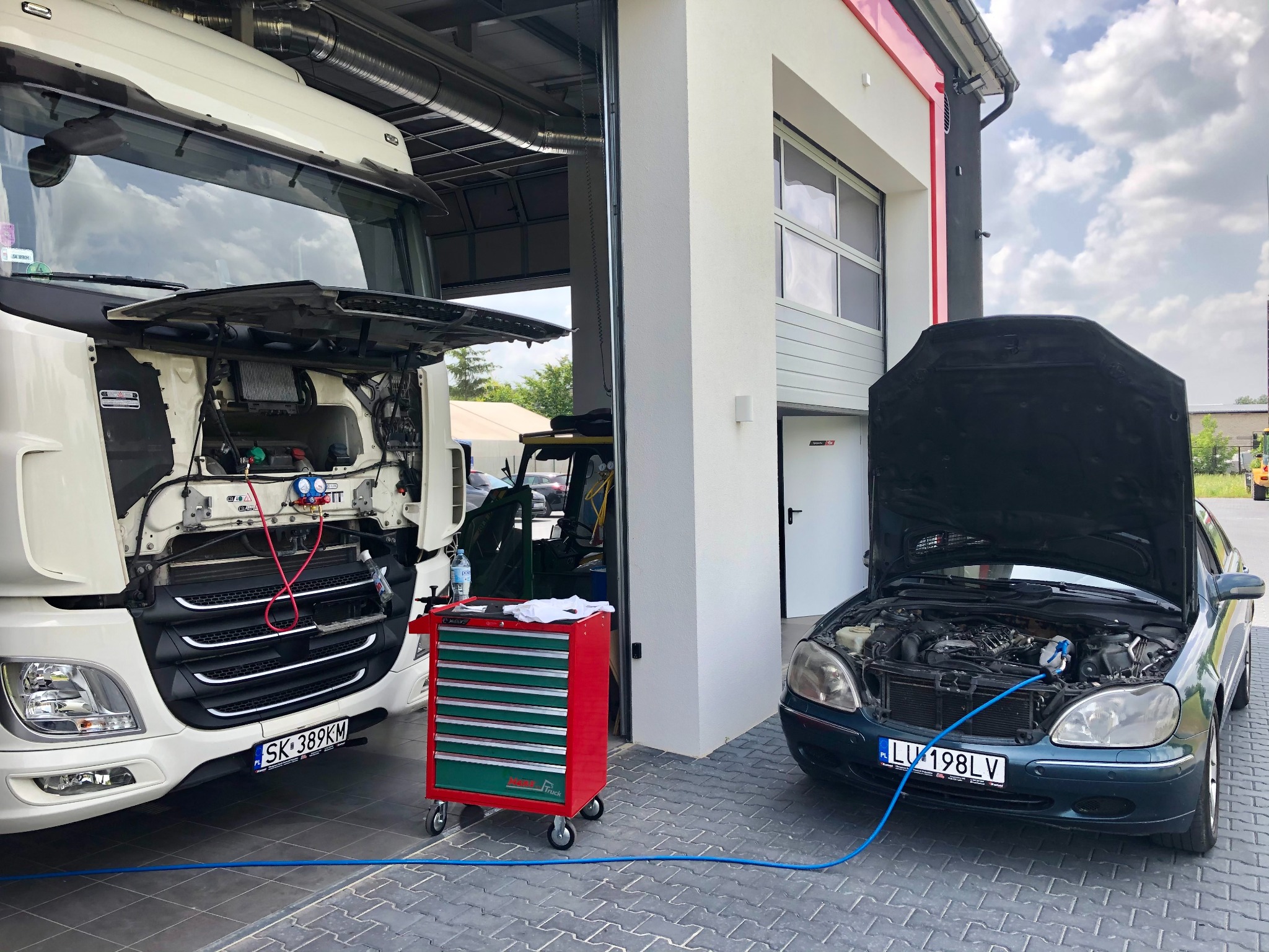 Serwis pojazdów: ciężarówka z otwartą maską i podłączonym sprzętem diagnostycznym obok ciemnego samochodu osobowego z otwartą maską, również z podłączonymi przewodami, na tle warsztatu.