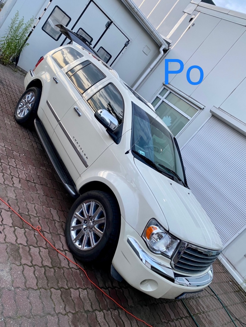 Biały Chrysler Durango po umyciu, zaparkowany na kostce brukowej przed budynkiem z otwartą klapą bagażnika, widoczny kabel na ziemi.