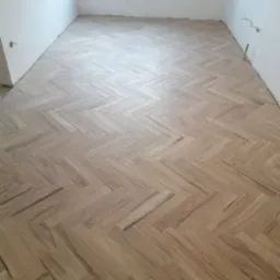 Jasne panele ułożone w jodełkę w pustym pomieszczeniu z białymi ścianami i widocznymi przewodami elektrycznymi.