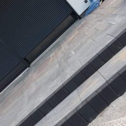 Choco - Ukośne ujęcie schodów zewnętrznych wyłożonych szarą kostką brukową z czarnymi obrzeżami, widoczne rolety okienne i fragment siatki zabezpieczającej.