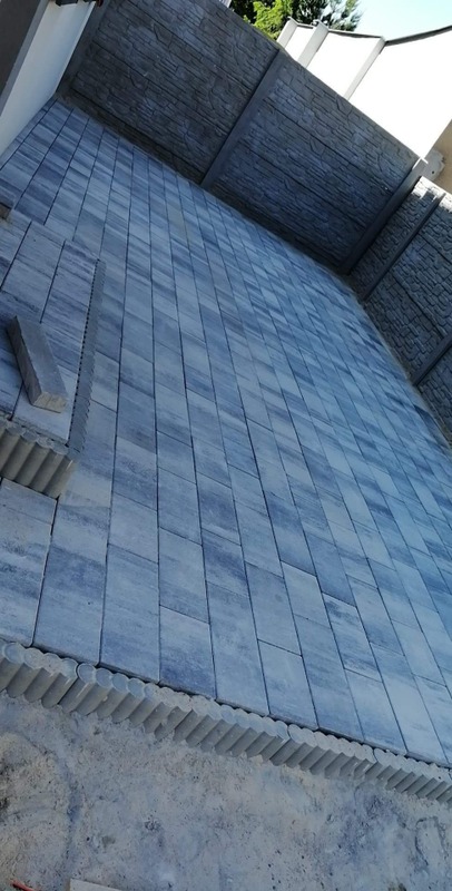 Niebiesko-szara kostka brukowa ułożona w regularny wzór, z obrzeżem z okrągłych elementów, widoczne szare betonowe panele ogrodzeniowe w tle, ujęcie z góry pod kątem.