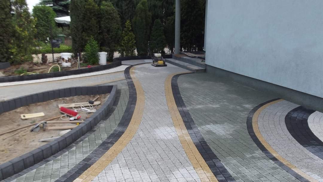 Ułożona z kostki brukowej ścieżka z żółtymi i czarnymi akcentami, prowadząca do schodów budynku, z zagęszczarką w tle i narzędziami brukarskimi po lewej stronie, otoczona zielenią.