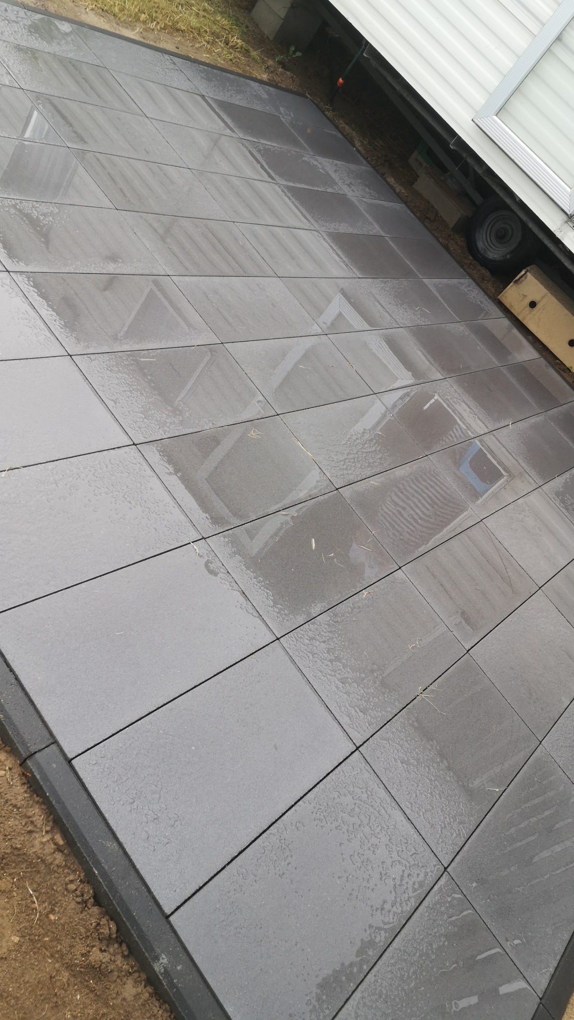Ułożona, mokra, szara kostka brukowa z ciemnoszarym obrzeżem, widoczny fragment przyczepy kempingowej w tle.