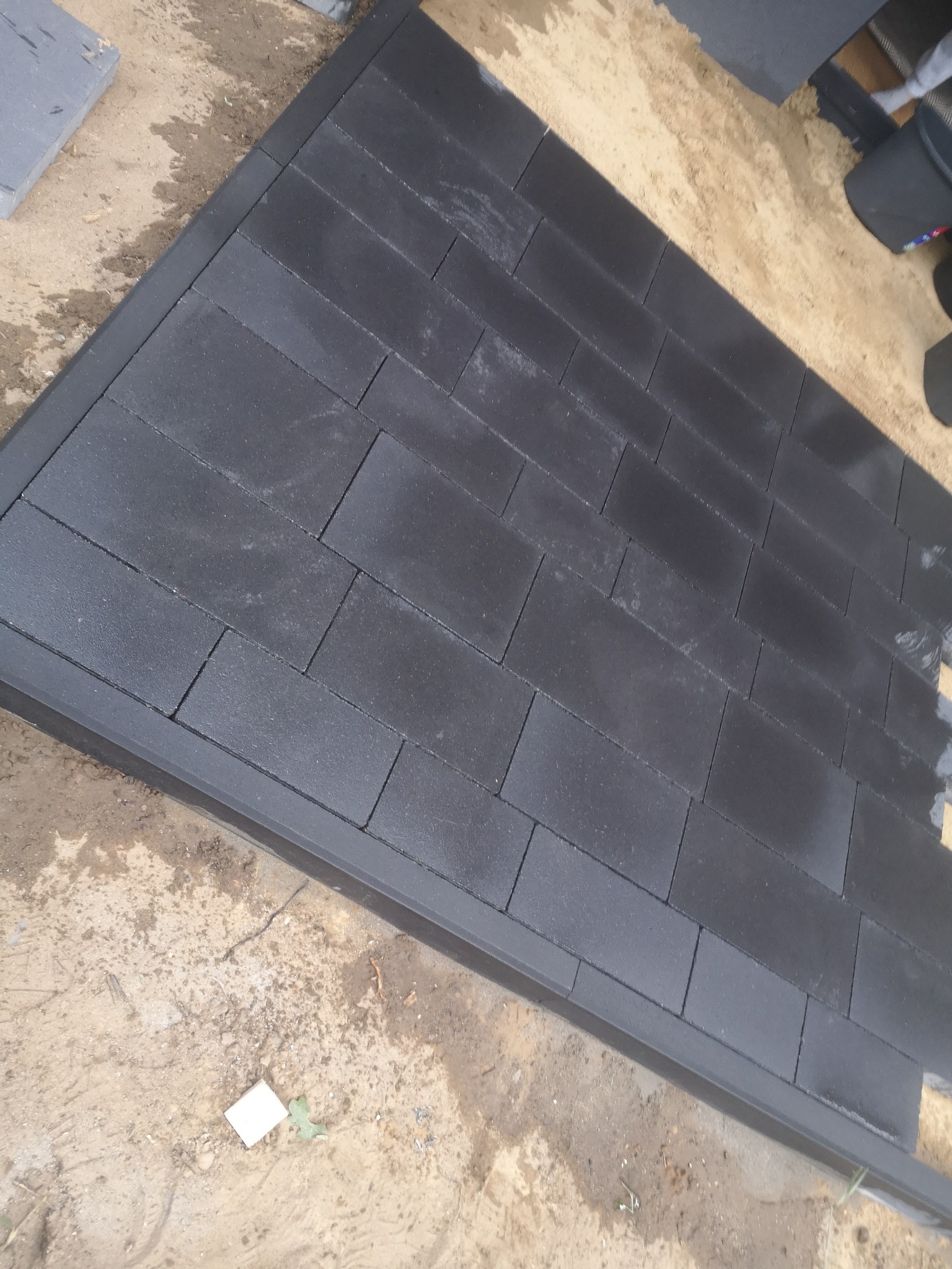 Ciemnoszara, prostokątna kostka ułożona w rzędach na piaskowym podłożu, obramowana ciemną obwódką. Widoczne ślady wilgoci na piasku.