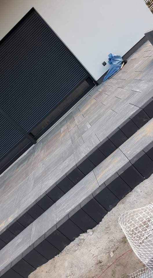 Ukośne ujęcie schodów zewnętrznych wyłożonych szarą kostką brukową z czarnymi obrzeżami, widoczne rolety okienne i fragment siatki zabezpieczającej.