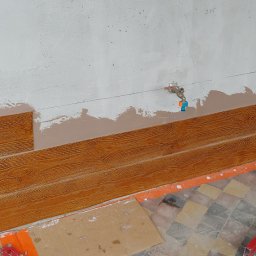 Paweł wojas - Montaż paneli ściennych imitujących drewno na nierównej ścianie, widoczna instalacja wodna i poziomica na podłodze w trakcie prac wykończeniowych.