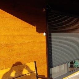 Paweł wojas - Zewnętrzna ściana domu z drewnopodobną elewacją w odcieniach brązu i zamontowaną szarą roletą okienną; widoczny fragment okna, fragment tarasu i zieleń w tle.