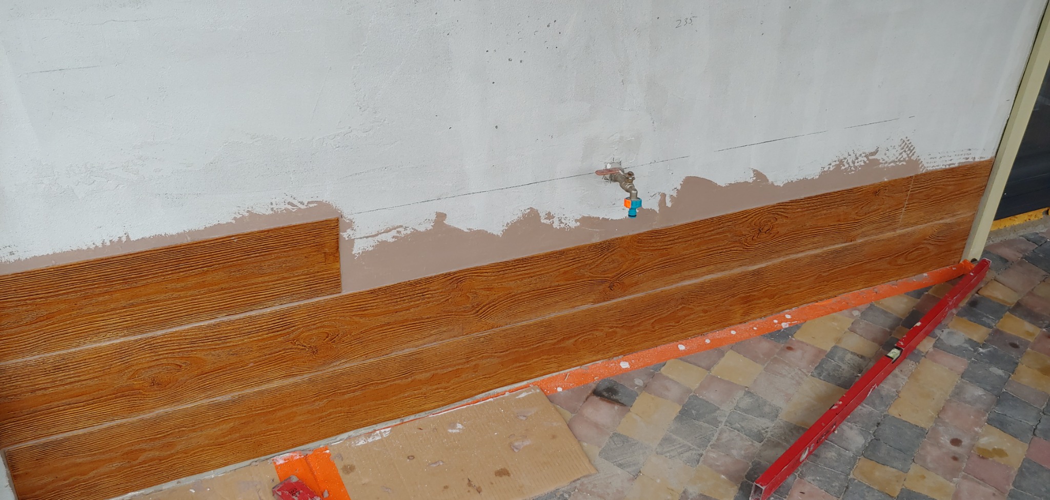 Montaż paneli ściennych imitujących drewno na nierównej ścianie, widoczna instalacja wodna i poziomica na podłodze w trakcie prac wykończeniowych.