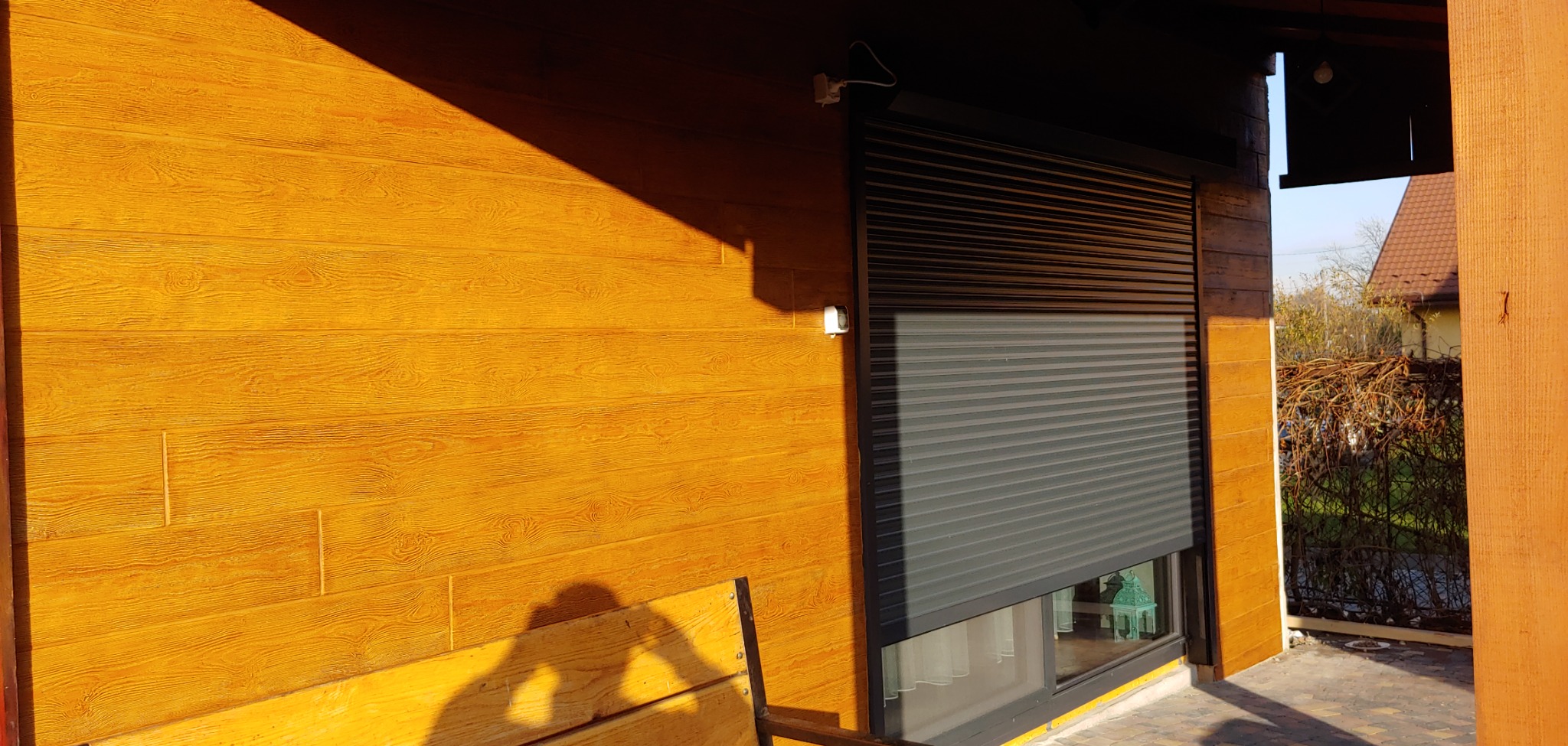 Zewnętrzna ściana domu z drewnopodobną elewacją w odcieniach brązu i zamontowaną szarą roletą okienną; widoczny fragment okna, fragment tarasu i zieleń w tle.