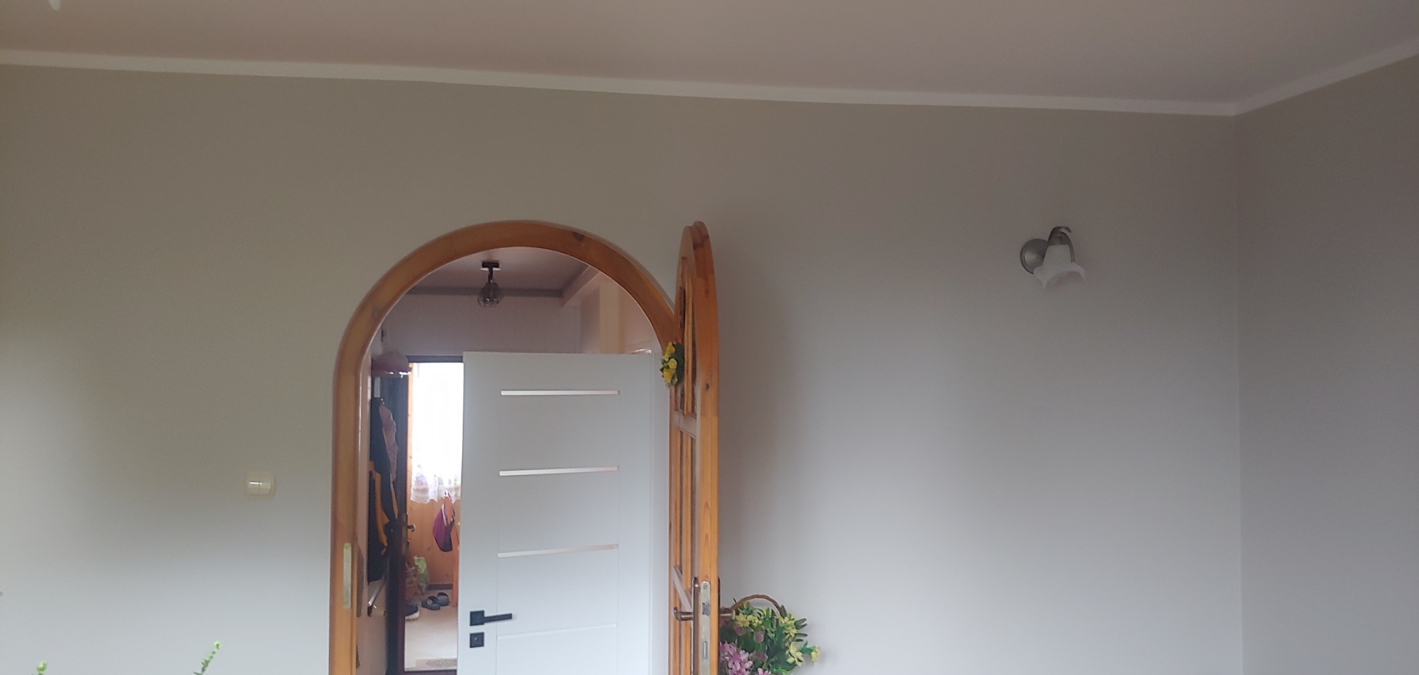 Pomalowane na szaro ściany z drewnianym łukiem drzwiowym i białymi drzwiami wewnętrznymi z poziomymi paskami, widoczny fragment sufitu i lampa, dekoracja kwiatowa na drzwiach i w koszyku.