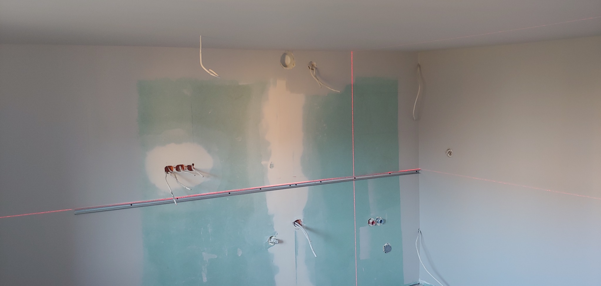Ściana w trakcie przygotowywania do prac wykończeniowych, widoczne płyty karton-gips z miejscami na gniazdka elektryczne i wyprowadzone przewody, poziomica laserowa wyznacza linię prostą.