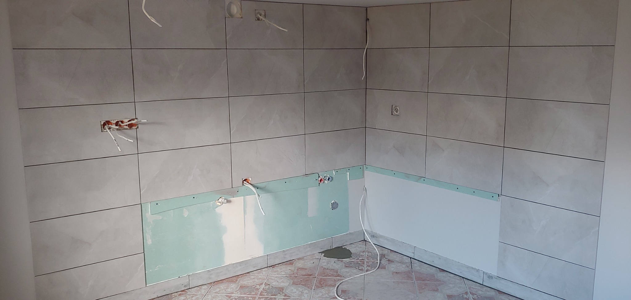 Ściana w trakcie układania płytek ceramicznych, widoczne wyprowadzone kable elektryczne, fragment płyty karton-gips i resztki zaprawy na podłodze.