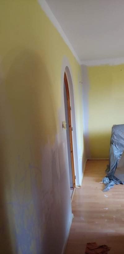 Żółta ściana w trakcie malowania z widocznymi śladami po gładzi, zabezpieczona podłoga i futryna drzwi łukowych, fragment sufitu z listwą dekoracyjną.