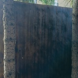 VANBUD - Ciemnobrązowe, drewniane ogrodzenie panelowe, częściowo przesłonięte przez pnie drzew, z widocznym fragmentem metalowej siatki i zabudowanej wiaty w tle.