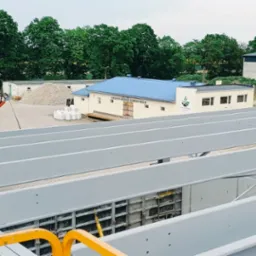 Szare, stalowe belki konstrukcyjne na tle budynków i zieleni. Widoczne szalunki fundamentowe. Kąt ujęcia z góry, podkreślający perspektywę. Dzień, światło rozproszone.