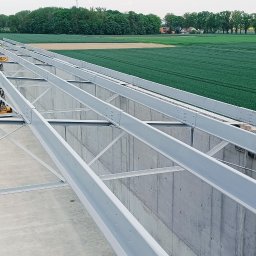 VANBUD - Konstrukcja stalowa hali w trakcie budowy, widok z góry na szkielet dachu. W tle zielone pola i las. Widoczne białe ściany i elementy konstrukcyjne.