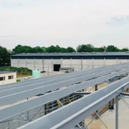 Konstrukcja stalowa hali w trakcie montażu, widok z góry. W tle budynek magazynowy i zieleń. Szara, pochmurna pogoda. Elementy konstrukcyjne pomalowane na szaro.