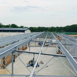 VANBUD - Wysoki kąt ujęcia szkieletu hali przemysłowej z betonu i stali na tle zielonych pól i budynków. Widoczna konstrukcja dachu i ściany. Prace w toku.