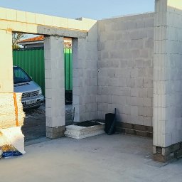 VANBUD - Wnętrze surowego budynku w trakcie budowy, z widocznymi ścianami z białych bloków i otworami drzwiowymi. Na zewnątrz zaparkowany srebrny samochód dostawczy.