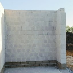 VANBUD - Wznoszone ściany z białych bloczków betonu komórkowego na fundamencie. Widoczny fragment terenu budowy i zieleń w tle. Kąt ujęcia z dołu podkreśla pionowość konstrukcji.