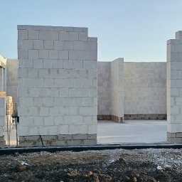 VANBUD - Wnętrze domu w trakcie budowy, ściany wzniesione z białych bloczków, widoczny fundament i teren budowy. W tle zaparkowane samochody i zieleń.