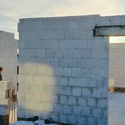 VANBUD - Wznoszenie ścian domu z białych bloczków, widoczny murarz przy pracy i częściowo wzniesiona konstrukcja w promieniach zachodzącego słońca.