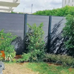 Nowoczesne ogrodzenie z paneli kompozytowych w kolorze grafitowym, częściowo przysłonięte przez bujną roślinność i kwiaty w ogrodzie.