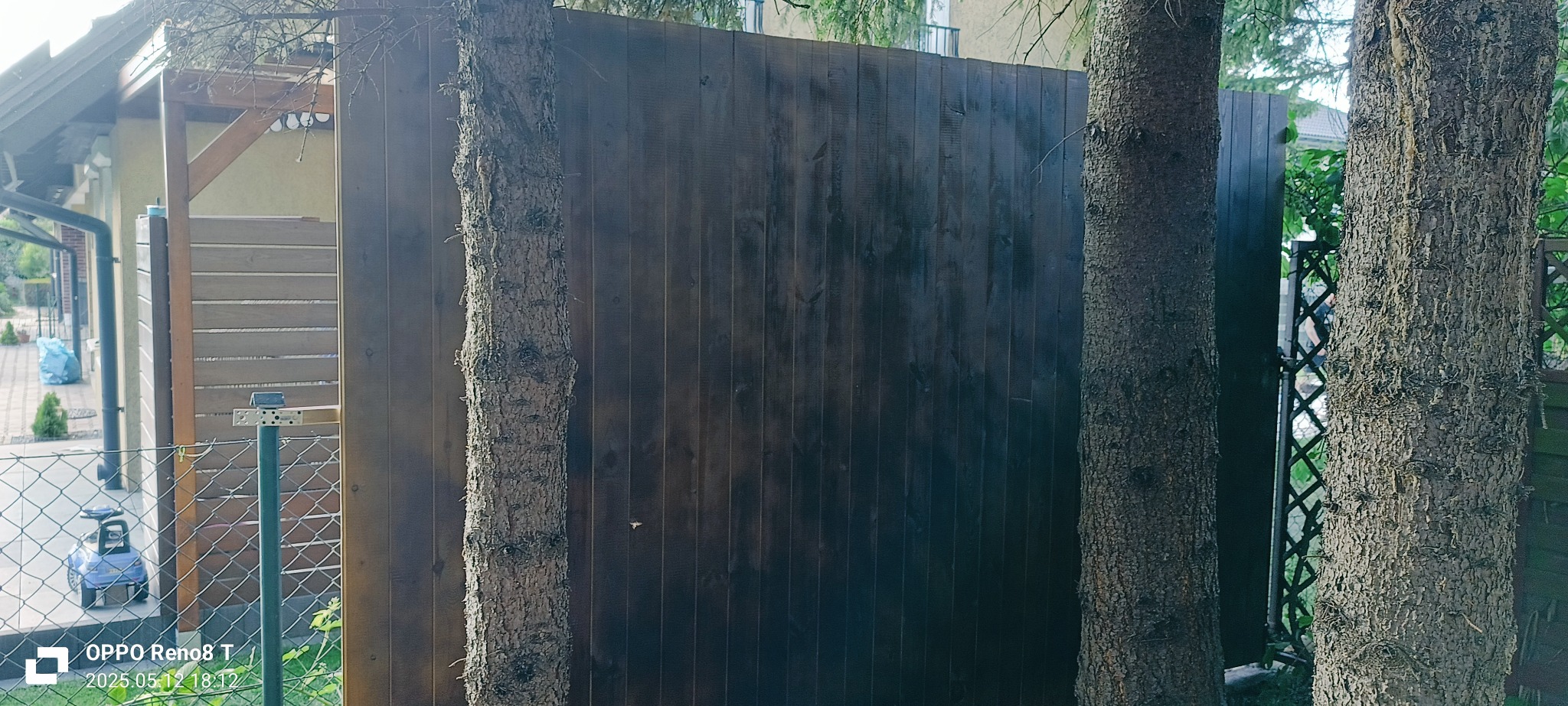 Ciemnobrązowe, drewniane ogrodzenie panelowe, częściowo przesłonięte przez pnie drzew, z widocznym fragmentem metalowej siatki i zabudowanej wiaty w tle.