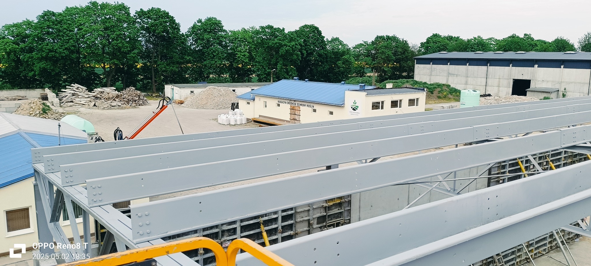 Szare, stalowe belki konstrukcyjne na tle budynków i zieleni. Widoczne szalunki fundamentowe. Kąt ujęcia z góry, podkreślający perspektywę. Dzień, światło rozproszone.